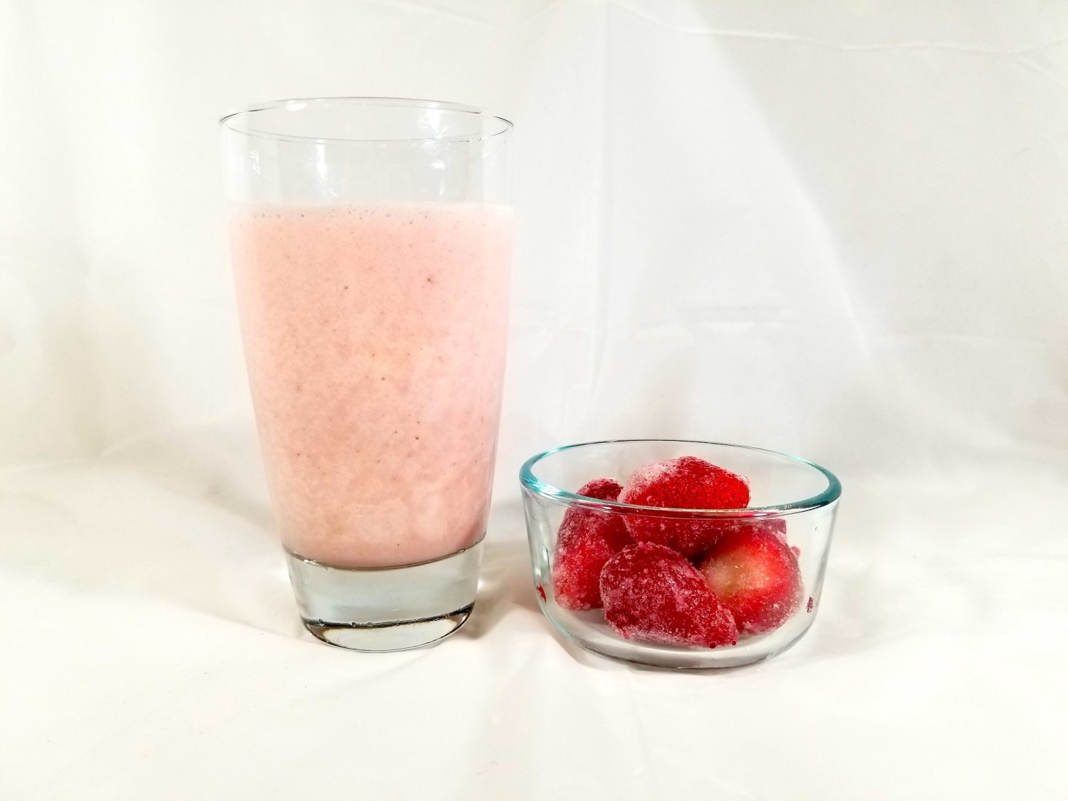 Strawberry bannana vanilla protein shake in a glass next to some frozen strawberries